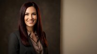 Professional businesswoman with long brown hair smiling confidently in dark blazer against neutral background for corporate marketing materials.