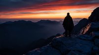 Silhouetted hiker stands on rocky mountain peak at dramatic sunset, gazing over misty valleys and distant ridges. Solitary adventurer contemplates expansive landscape during golden hour.