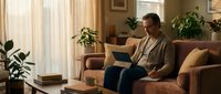 Middle-aged Caucasian man working on laptop while sitting on brown leather sofa in warm, plant-filled living room with natural light from windows.
