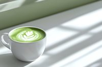 Green matcha latte with artistic foam heart design in white ceramic cup on light surface with diagonal window shadows creating minimalist composition.