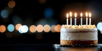 Birthday cake with glowing candles on dark background with bokeh lights, festive celebration dessert with white frosting and golden crust.