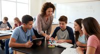 Female teacher with curly hair helping diverse group of teenage students with tablet and worksheets in bright modern classroom setting.