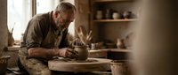 Senior male potter shaping clay on spinning wheel in traditional ceramic workshop with natural lighting and rustic shelving displaying handmade pottery pieces.