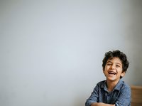 Young boy with dark curly hair and olive complexion, wearing blue checkered shirt, smiling broadly against white wall. Suitable for educational materials, family content, and lifestyle publications.