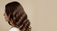 Woman with long, voluminous wavy brown hair styled in loose waves, shown in profile against a neutral beige background. Suitable for hair care, beauty, and salon marketing.