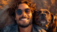 Young Hispanic man with curly hair and sunglasses smiling while lying next to his brown dog outdoors during golden hour sunset, capturing joyful friendship moment.