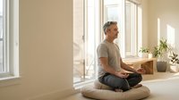 Middle-aged Caucasian man practicing meditation in bright modern room with large windows, sitting cross-legged on cushion in peaceful morning light.