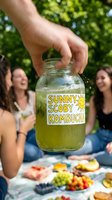 Hand holding a glass jar of green kombucha at a sunny outdoor picnic with friends laughing in the background, fresh fruit spread visible.