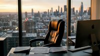 Executive leather office chair at modern workplace desk with city skyline view through floor-to-ceiling windows during golden hour sunset creating professional atmosphere.