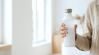Woman holding white reusable water bottle with metal cap in bright interior space, promoting sustainable hydration and eco-friendly lifestyle choices.