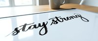 Stay strong handwritten text on white paper with coffee cup and notebook on wooden desk, motivational message for resilience and determination.