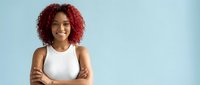 Confident young African American woman with curly red hair smiling with arms crossed on light blue background, isolated for business and lifestyle designs.
