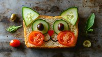 Creative food art featuring toast decorated as cat face with cucumber ears, olive eyes, cherry tomato cheeks, and fresh herbs on rustic background.