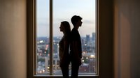 Silhouette of young couple standing back to back by large window overlooking city skyline at dusk. Relationship conflict and communication problems concept.