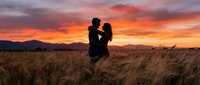 Romantic couple silhouette embracing in golden wheat field at sunset with dramatic orange sky and mountain backdrop for wedding and love concepts.