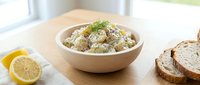 Creamy potato salad with fresh herbs served in white bowl on wooden table with lemon and bread slices for lunch or dinner meal preparation.