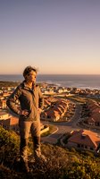 Middle-aged Caucasian male in casual athletic wear standing on hillside overlooking coastal suburban neighborhood at golden hour sunset, contemplating ocean view for lifestyle and travel concepts.