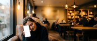 Young woman with dark hair resting head on hand while holding smartphone in cozy coffee shop with warm lighting and blurred customers in background.