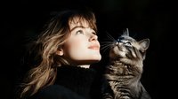 Young Caucasian woman in black turtleneck with long brown hair gazing upward alongside striped tabby cat against dark background, creating emotional bond moment.