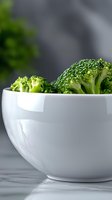 Fresh green broccoli florets in white ceramic bowl on marble surface against dark background, minimalist food styling for healthy eating concept.