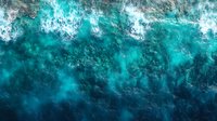 Aerial view of turquoise ocean waves crashing with white foam creating dynamic patterns. Deep blue tropical waters photographed from above showing natural movement.