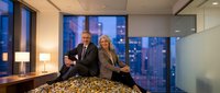 Confident business partners sitting on desk in modern office with city skyline view during evening hours, representing successful corporate teamwork.