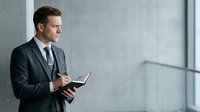 Caucasian male professional in dark business suit and tie holding tablet device against modern gray concrete wall, suitable for corporate, technology, and business concepts.