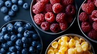 Fresh ripe berries in metal bowls on dark background. Colorful mix of blueberries, raspberries and yellow raspberries arranged in modern composition.