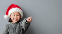 Cheerful Asian girl in Santa hat pointing with excitement, wearing cozy gray sweater against neutral background. Perfect for holiday advertising.