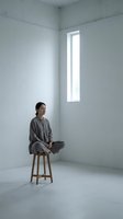 Young Asian woman in gray clothing meditating on wooden stool in minimalist white room with natural light from narrow window for wellness and mindfulness concepts.