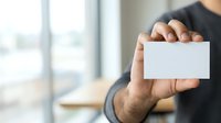 Man in black shirt holding blank white business card in office environment with window background.