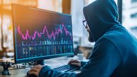 Young male trader in hoodie analyzing financial market data and stock charts on computer monitor in modern office with evening sunlight through window.