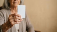 Senior woman with gray hair and wrinkled hands holding blank white card for voting, election, or message display against neutral beige background.