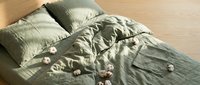 Unmade bed with rumpled olive green linen bedding and scattered white flowers on wooden floor in bright natural sunlight creating cozy morning atmosphere.