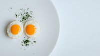 Two sunny side up fried eggs with bright orange yolks garnished with fresh herbs on minimal white plate against light background, food photography.