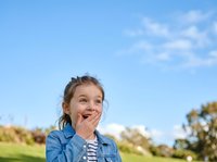 Young girl with brown hair and light skin, wearing a denim shirt over striped top, smiling with hand to mouth in outdoor park setting with green grass and blue sky.