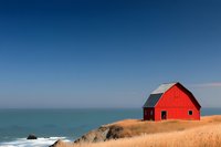 Red barn on coastal cliff overlooking turquoise ocean waters, golden dry grass in foreground against deep blue sky, dramatic seaside landscape.