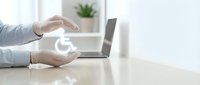 Hands protecting wheelchair accessibility symbol on desk with laptop in modern office environment promoting disability inclusion and equal opportunities.