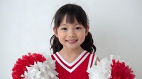 Young Asian girl with pigtails wearing red and white cheerleader uniform holding pom-poms against light background, smiling brightly for school spirit portrait.