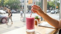 A person with medium skin tone holds a straw in a vibrant pink smoothie drink served in a glass mug, positioned on a wooden table by a window overlooking an urban street scene with pedestrians and vehicles.