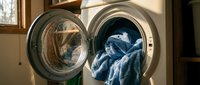 Modern white washing machine with open door showing blue denim clothes inside, warm sunlight streaming through laundry room creating cozy domestic atmosphere.
