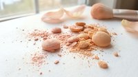 Crushed and whole makeup powder in warm peachy and nude tones scattered on white surface with brushes, suitable for beauty, cosmetics, and skincare product advertising.