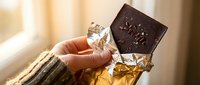 Mature woman holding crystal glass of whiskey with dark chocolate bar, warm sunlight streaming through window, luxury lifestyle and indulgence concept for food and beverage marketing.