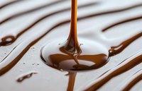Liquid chocolate dripping and forming glossy puddle on white surface, close-up view showing rich texture and smooth flow of melted dark chocolate.