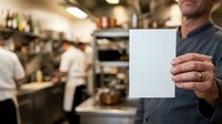 Middle-aged male chef in professional kitchen holding blank white card for menu or promotional text, with blurred staff and cooking equipment in background.