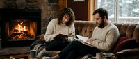 Cozy couple reading and writing in notebooks by a warm fireplace in a rustic cabin living room during winter, relaxing together indoors.