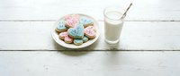 Colorful decorated sugar cookies with pastel icing on white plate next to glass of milk on rustic wooden table for sweet treats and baking concepts.