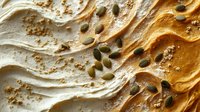 Creamy peanut butter swirl texture with scattered green pumpkin seeds creating dynamic waves and patterns, macro food photography on light background.