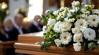 Elegant white floral arrangement on wooden casket at funeral ceremony with mourners in background paying respects.