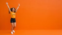 Young woman with curly hair, medium skin tone, wearing yellow crop top and black shorts, jumping with arms raised in celebration against vibrant orange background.